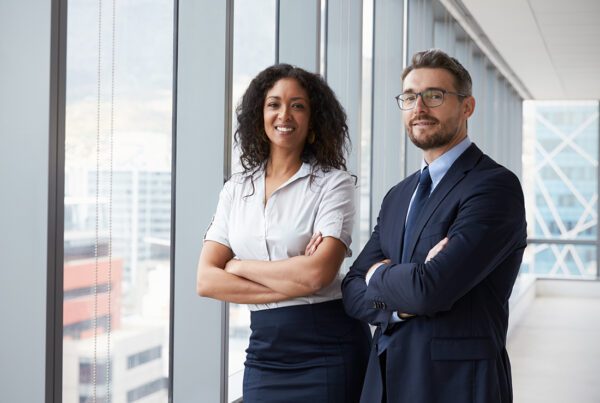 Business Insurance - Business Man and Woman Stand Together With Their Arms Crossed and Smiling at an Office With Large Glass Windows
