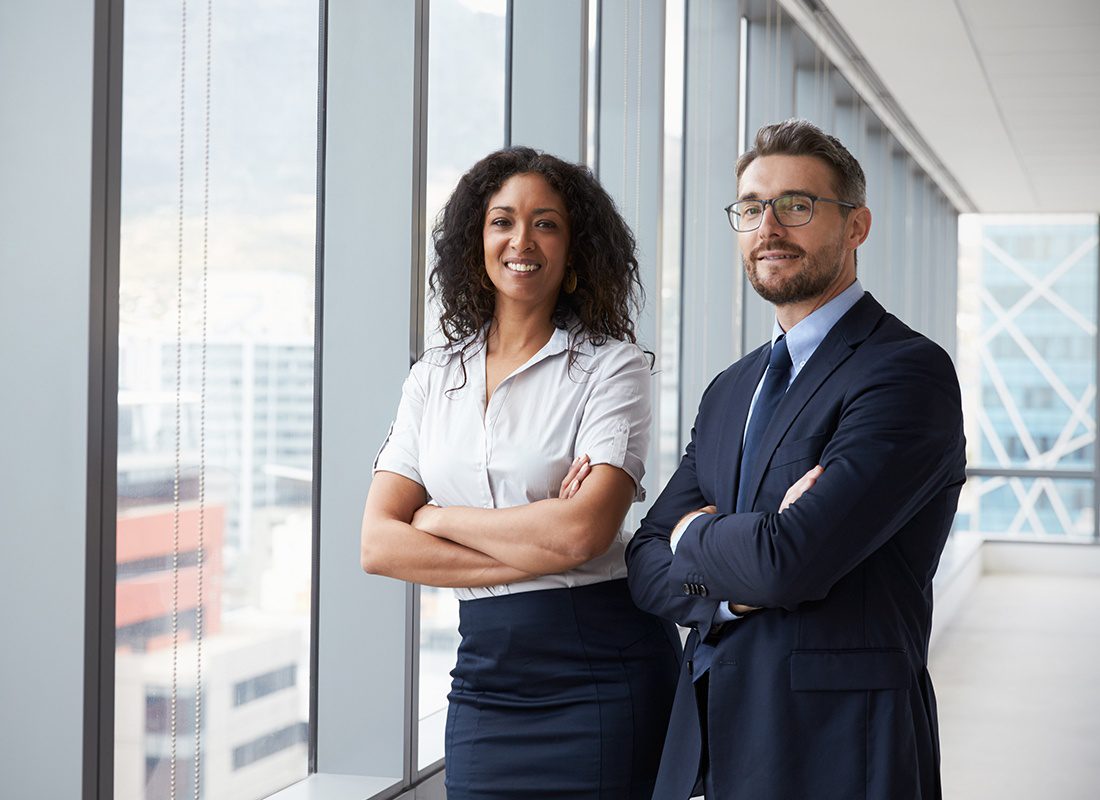 Business Insurance - Business Man and Woman Stand Together With Their Arms Crossed and Smiling at an Office With Large Glass Windows