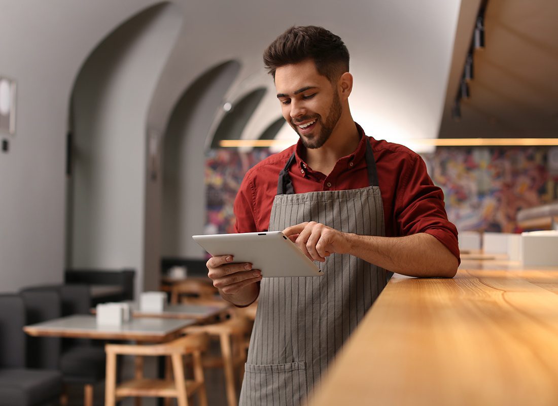 Service Center - Smiling Business Owner Wearing an Apron Leans Against a Counter While Using a Tablet at His Cafe