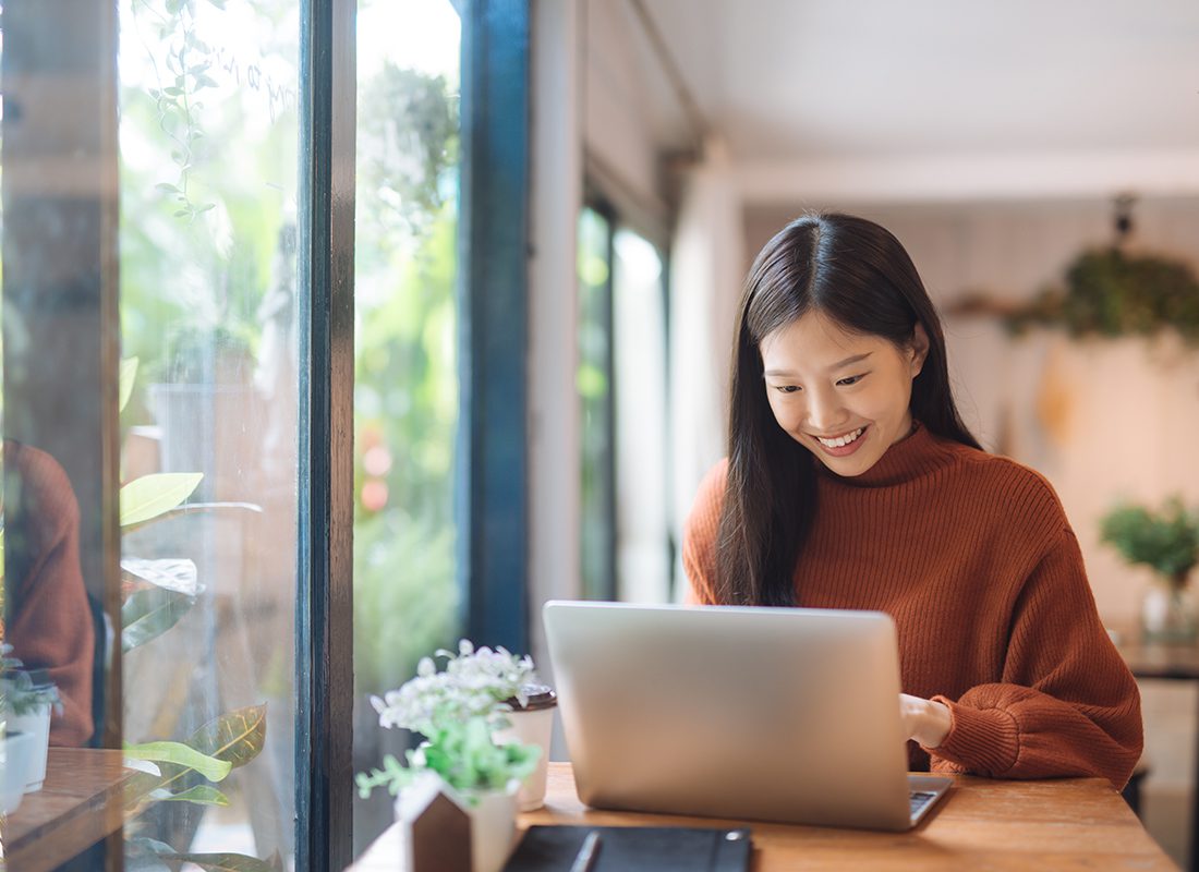 Blog - Young Woman Smiling Down at Her Computer As She Sits at a Wooden Table Next to a Large Window on a Nice Day